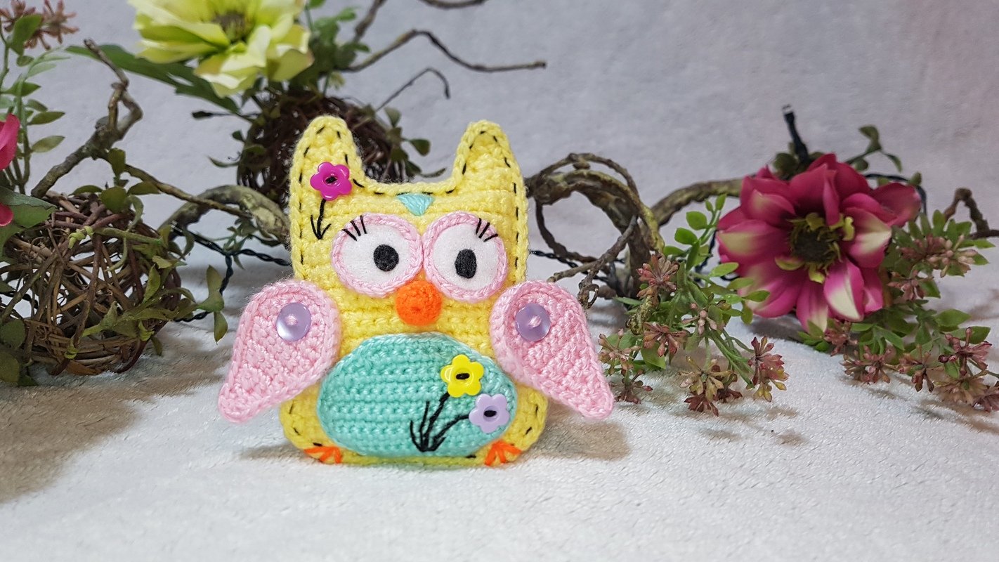 Gehäkelte gelbe Amigurumi-Eule mit rosa Flügeln und mintgrünem Bauch vor Dekozweigen