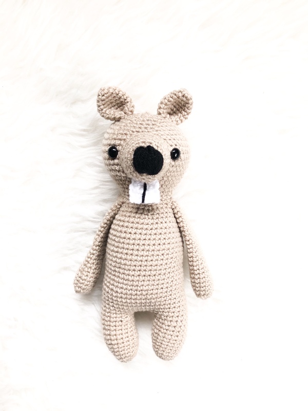 Beiger gehäkelter Amigurumi-Biber mit schwarzen Augen und zwei weißen Vorderzähnen, liegend auf hellem Untergrund