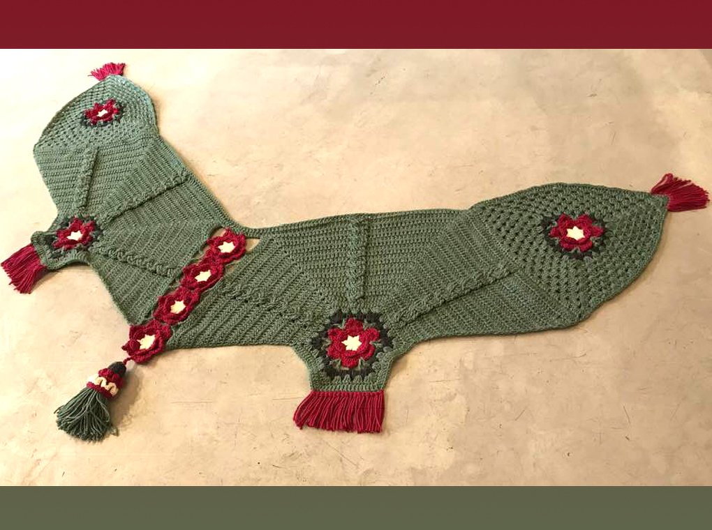 Grünes gehäkeltes Tuch mit roten Blütenapplikationen und Quasten auf hellem Untergrund.