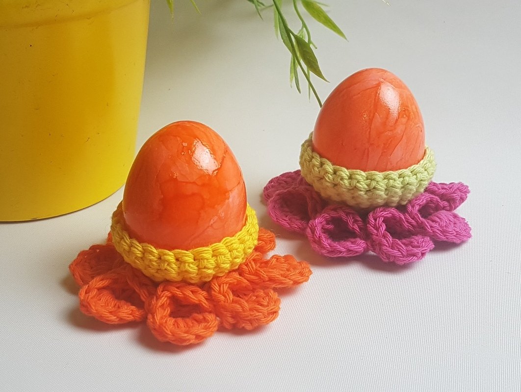 Zwei orangefarbene Eier in gehäkelten Blumen-Eierbechern, einer orange mit gelbem Saum, einer pink mit hellgrünem Saum