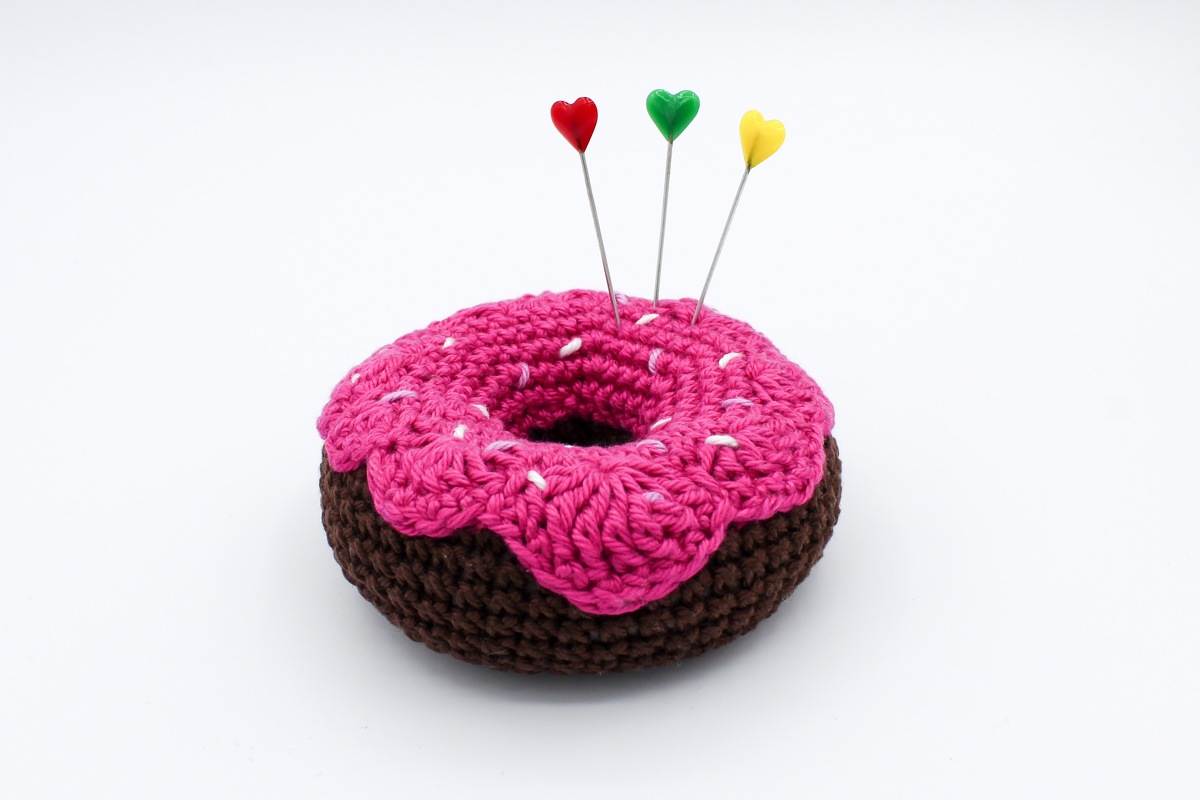 Gehäkeltes Donut-Nadelkissen, braune Basis mit pinker Glasur und drei herzförmigen Stecknadeln