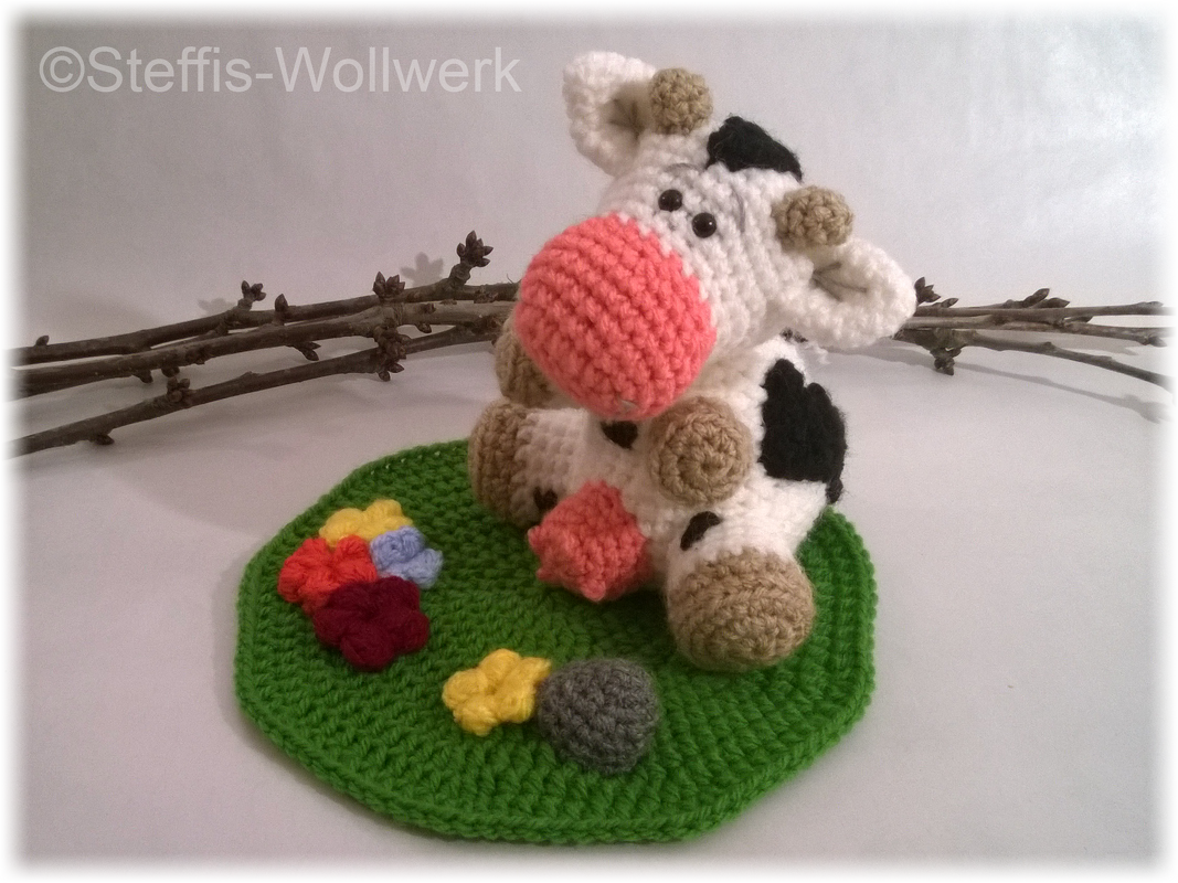 Gehäkelte Amigurumi-Kuh in Schwarz-Weiß mit rosa Schnauze auf grüner Häkelwiese mit kleinen Blüten