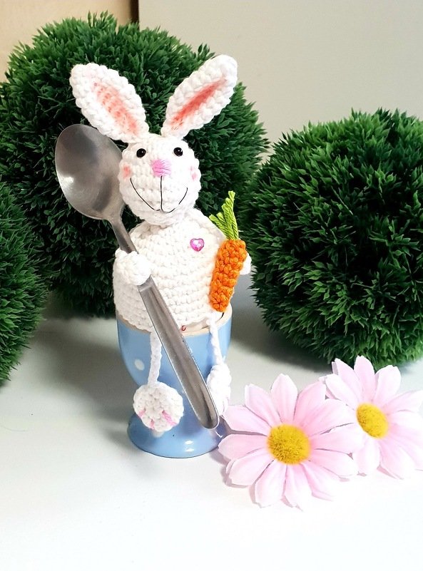 Weiß gehäkelter Osterhase als Eierwärmer in blauem Eierbecher, hält einen Löffel und eine kleine Karotte, vor grüner Deko und rosa Blumen.