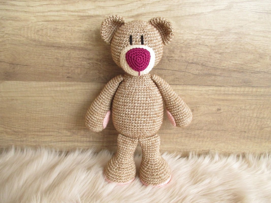 Gehäkelter beiger Amigurumi-Teddy mit rosa Nase auf hellem Fell vor Holz