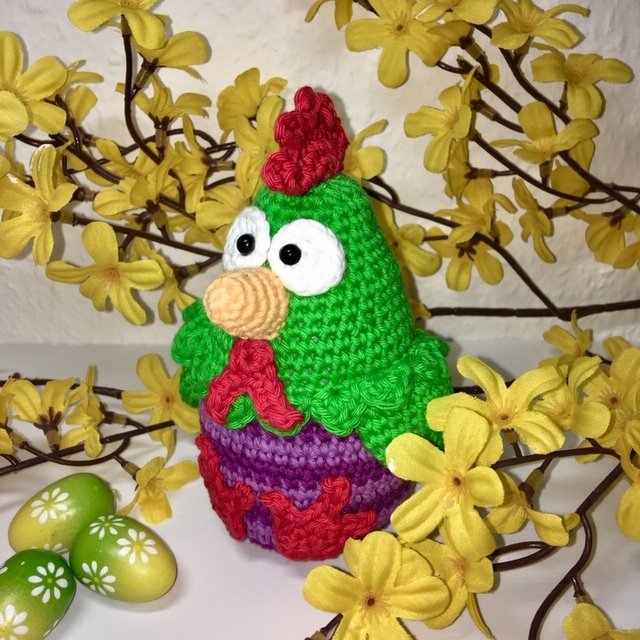 Gehäkeltes Amigurumi-Huhn in Grün und Lila vor gelben Blütenzweigen und grünen Ostereiern.