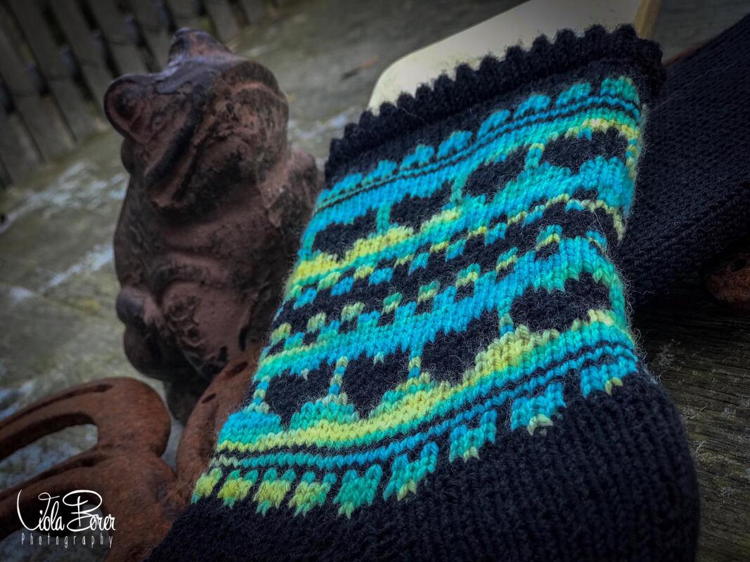 Nahaufnahme einer handgestrickten Socke mit Herzmuster in Türkis und Lindgrün auf schwarzem Grund, daneben eine kleine Froschfigur.