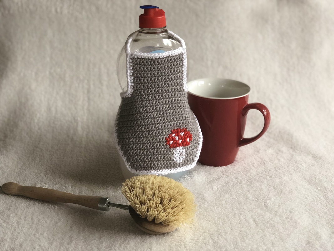 Gehäkelte Hülle für eine Spülmittelflasche, grau mit rotem Pilz, daneben rote Tasse und Spülbürste.