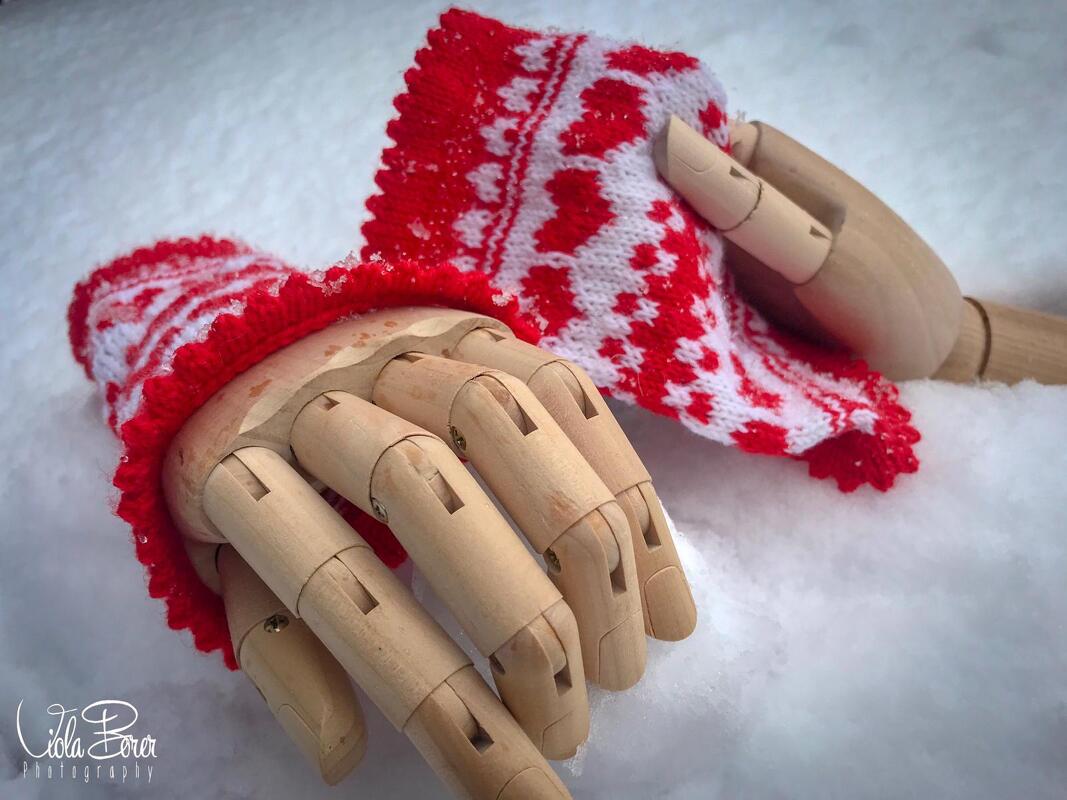 Hölzerne Gliederhände mit gestrickten roten und weißen Armstulpen mit Herzmuster auf Schnee.