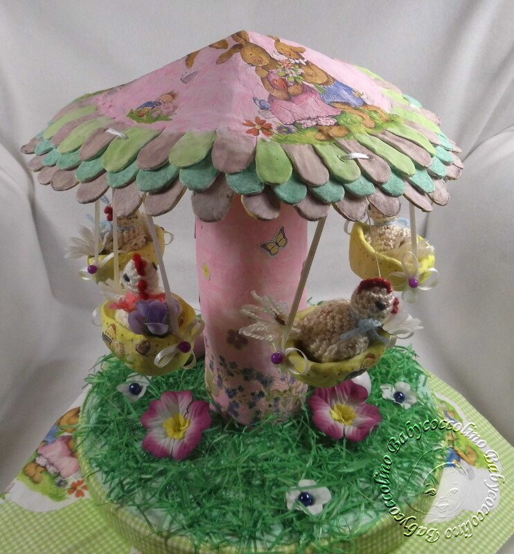 Rosa-grünes Bastelkarussell mit Hasen auf dem Dach und kleinen Kükenfiguren in Schaukeln über einer grünen Grasfläche.