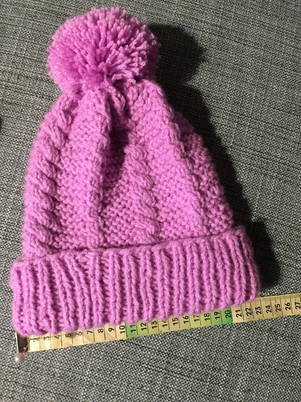 Fliederfarbene Strickmütze mit Zopfmuster, Rippenbund und Bommel auf grauem Stoff neben einem Maßband.