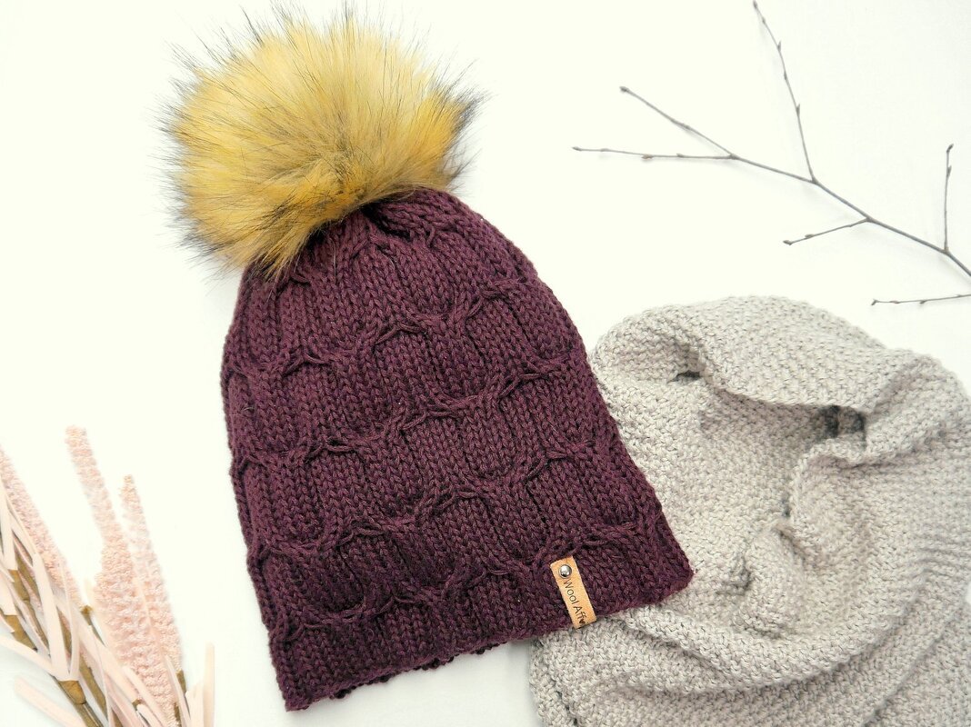 Dunkellila gestrickte Beanie mit großem gelblich-braunem Bommel neben hellgrauem Schal