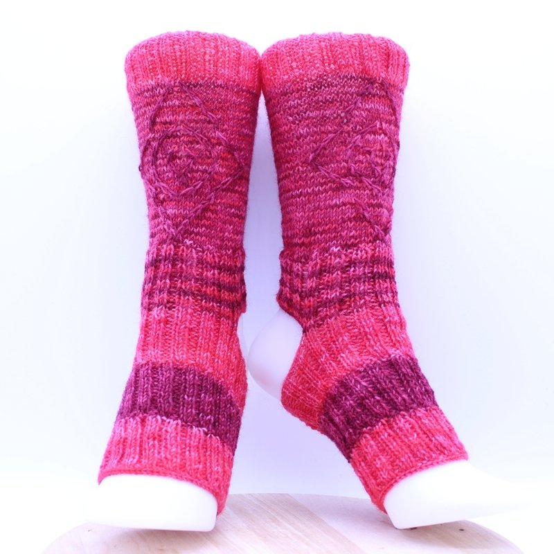 Zwei gestrickte Beinstulpen in Pink- und Rottönen mit Herz-Zopfmuster auf weißen Fußformen präsentiert.