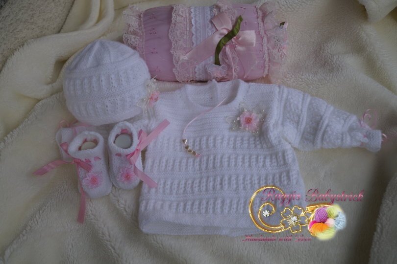 Weißes gestricktes Baby-Set mit Pullover, Mütze und Schühchen, rosa Schleifen und Blumenverzierung auf cremefarbener Decke