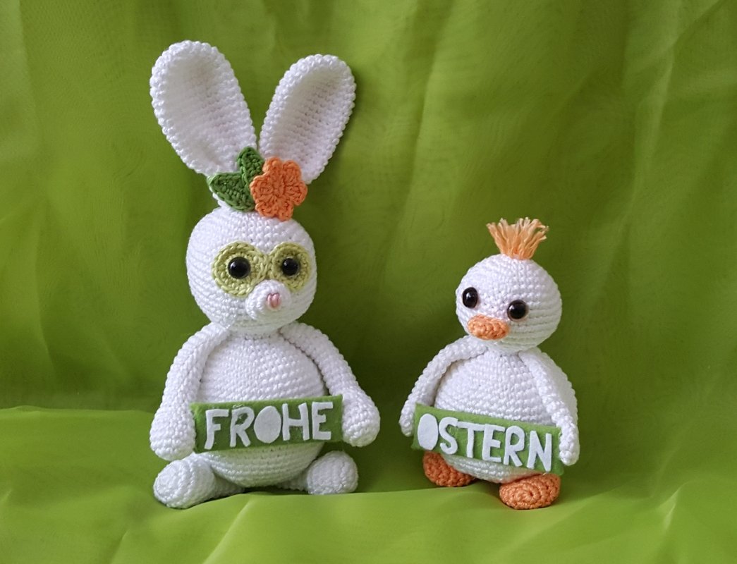 Zwei weiße gehäkelte Amigurumi-Figuren, Hase mit langen Ohren und Küken, halten grüne Schilder 'FROHE OSTERN' vor grünem Hintergrund