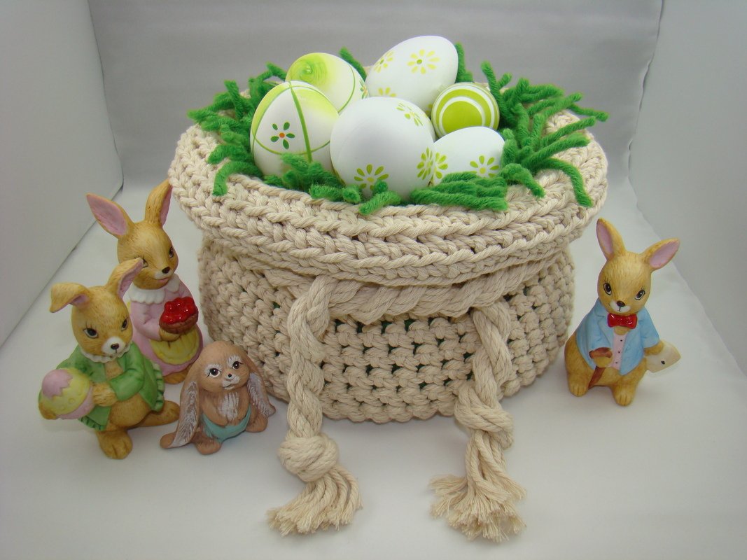 Beigefarbener gehäkelter Korb mit Kordelzug, gefüllt mit weiß-grünen Ostereiern auf grünem Garn, umgeben von Hasenfiguren