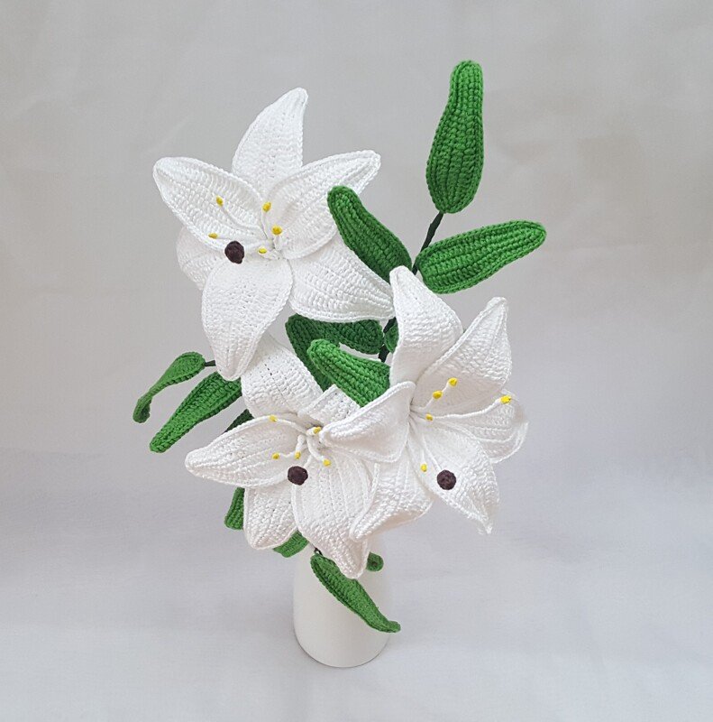 Three white crocheted lily flowers with green leaves in a white vase