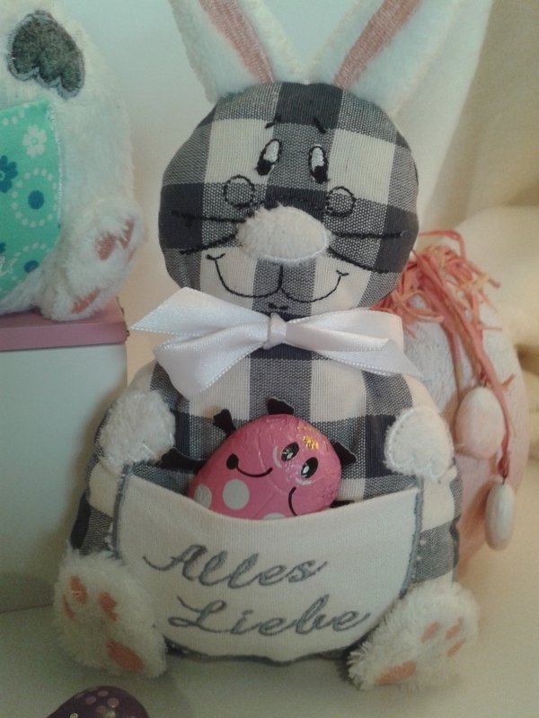 Kariertes Stoffhäschen (Vichy-Karo) mit weißer Schleife und aufgesetzter Tasche 'Alles Liebe', hält ein rosa Schokoladenei