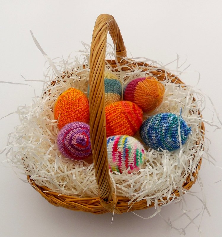 Gestrickte Ostereier mit verschiedenen Mustern und Farben in einem Weidenkorb auf Papierstreu