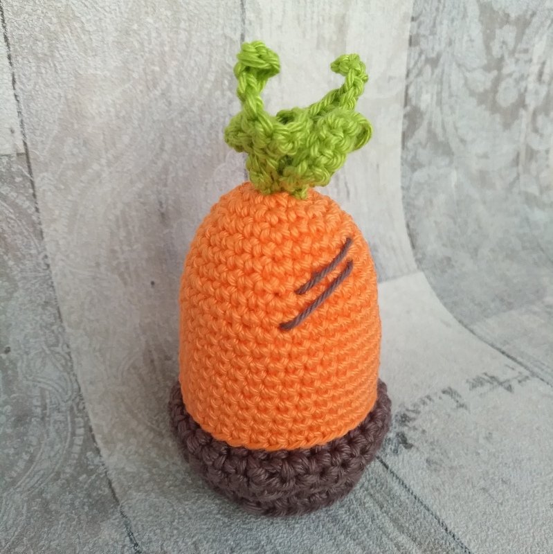 Gehäkelter Karotten-Eierwärmer mit grünen Blättern und braunem Sockel auf hellem Hintergrund.