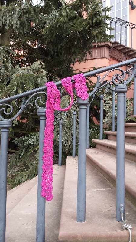 Gehäkelter, langer pinker Schal mit Spitzenmuster über einem dunklen Metallgeländer an einer Außentreppe
