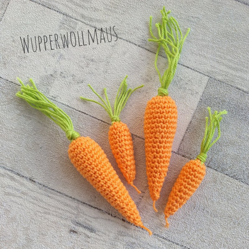 Vier gehäkelte orangefarbene Karotten mit grünen Garnblättern in verschiedenen Größen auf hellem Untergrund