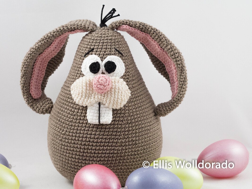 Gehäkelter brauner Hase mit rosa Innenohren und weißen Zähnen neben bunten Ostereiern auf hellem Hintergrund.