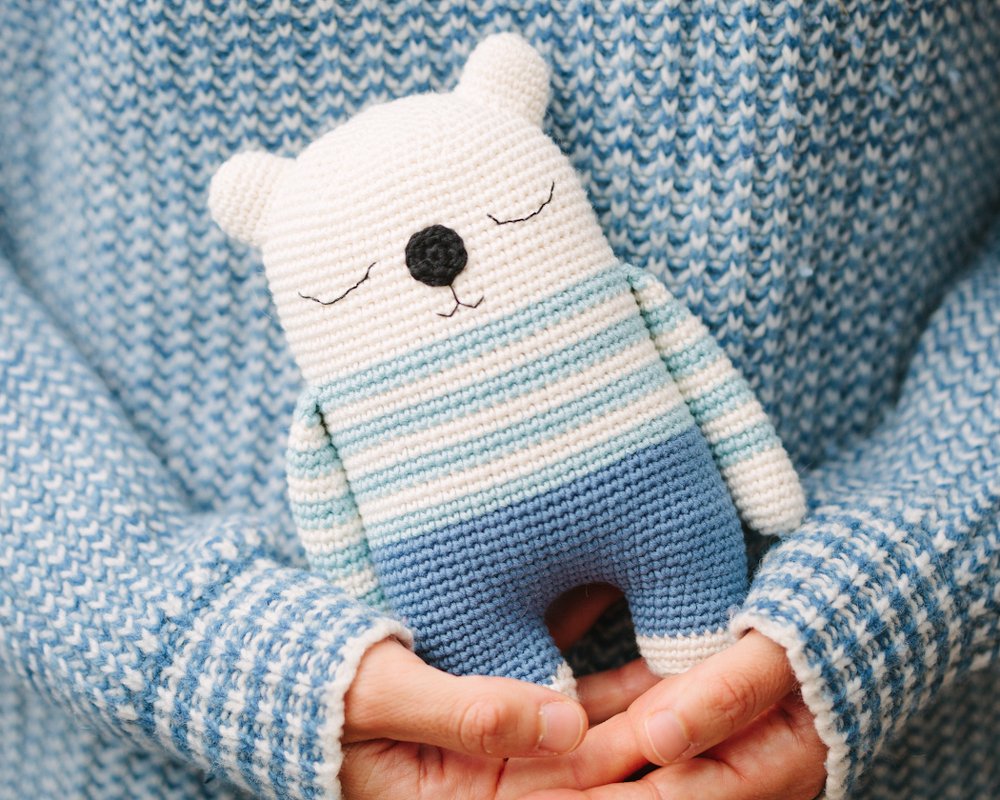crocheted amigurumi teddy bear with blue and white stripes, embroidered eyes and nose, held in hands