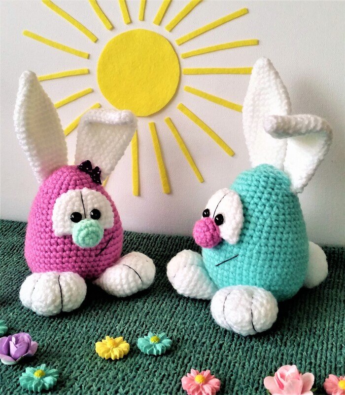 Zwei gehäkelte Amigurumi-Hasen in Rosa und Mint mit weißen Ohren und Pfoten vor einer gelben Sonnendekoration