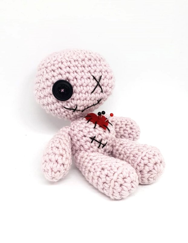 Rosa gehäkelte Amigurumi‑Puppe im Voodoo‑Stil mit schwarzem Knopfauge, gesticktem X‑Auge, rotem Filzherz und Stecknadeln auf weißem Hintergrund.