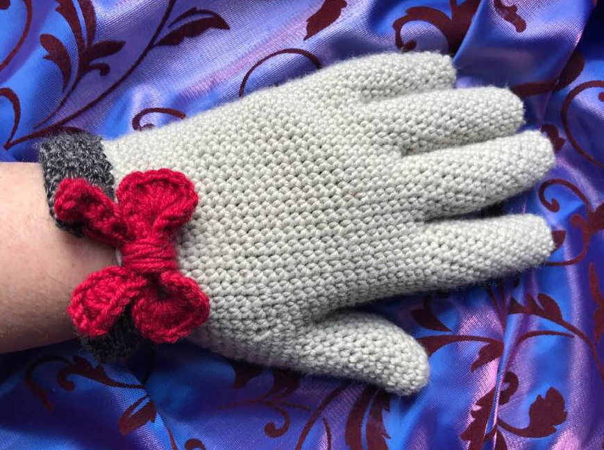 Hellgrauer, gehäkelter Damenhandschuh mit roter Schleife auf violettem, gemustertem Stoff
