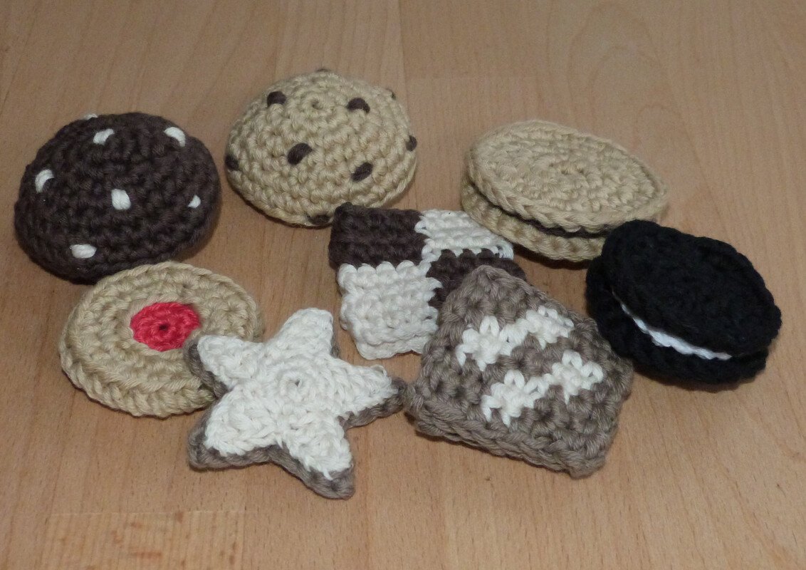 Acht gehäkelte Spielzeugkekse in unterschiedlichen Formen und Farben auf hellem Holzboden angeordnet.