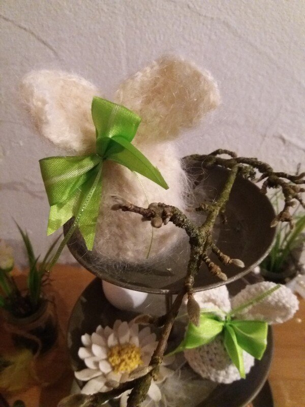 Zwei gehäkelte Häschen-Eierwärmer aus flauschigem Garn mit grünen Schleifen auf einer Etagère neben Zweig und Blüte