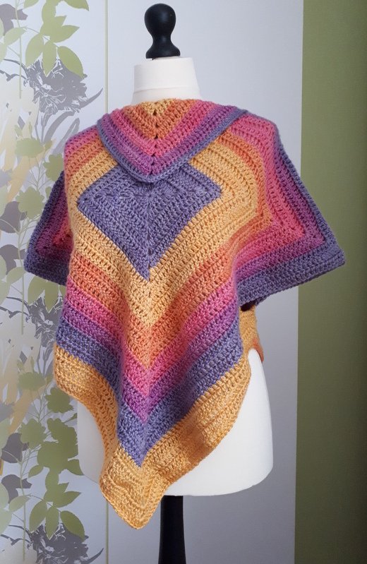 Gehäkelter Poncho in Lila, Pink, Orange und Gelb mit Rautenmuster auf einer Schneiderpuppe.
