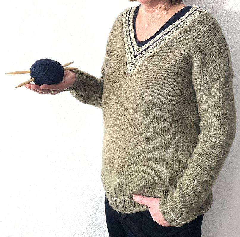 Überschnittener V‑Pullover in Oliv, glatt gestrickt, mit Rippenbündchen; Garnknäuel und Stricknadeln in der Hand