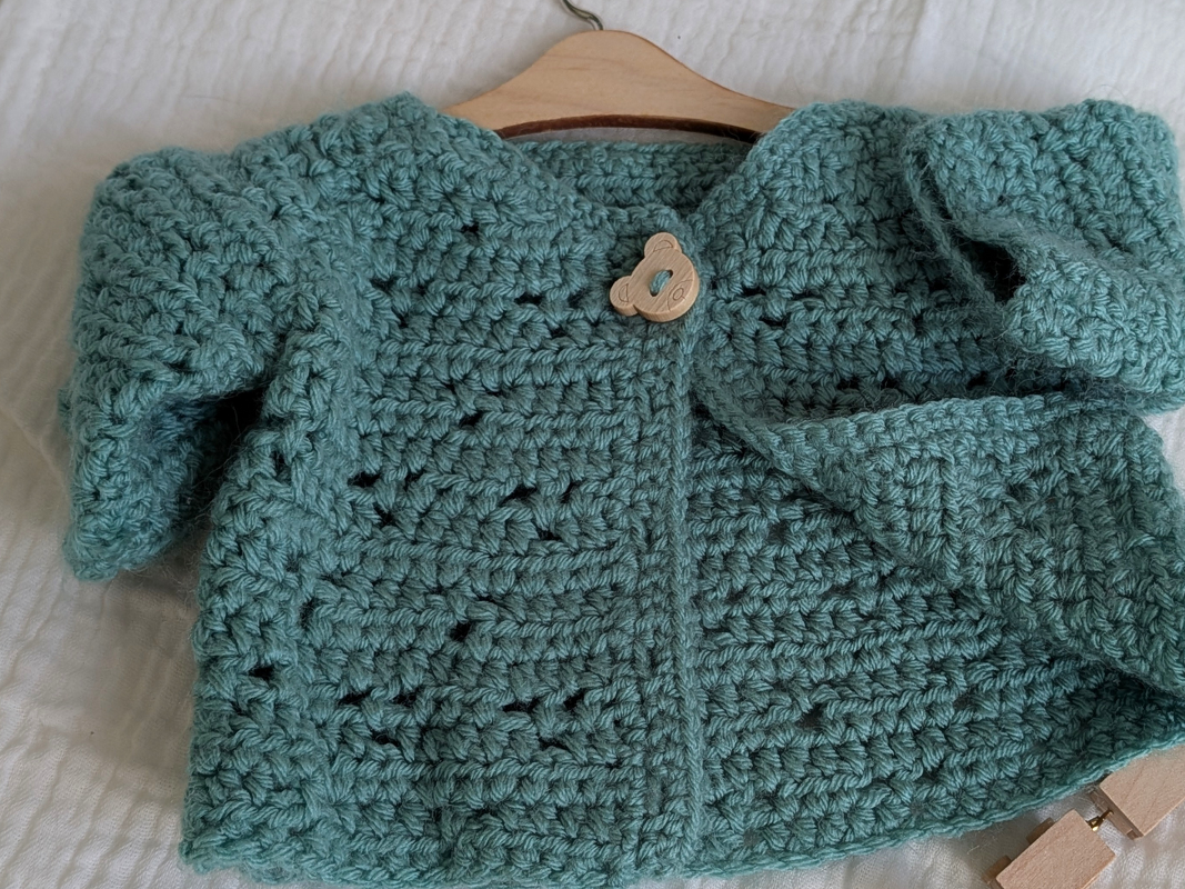 Türkise gehäkelte Babyjacke mit Holzknopf in Bärenform auf Holzbügel
