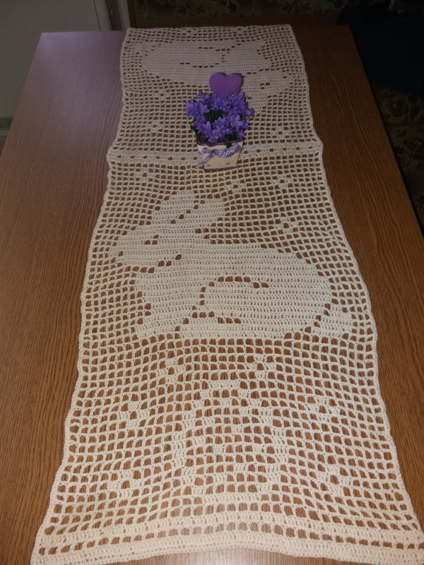 Beigefarbener gehäkelter Tischläufer mit Hasenmotiven auf einem Holztisch, dekoriert mit lila Blumen in einem kleinen Topf.