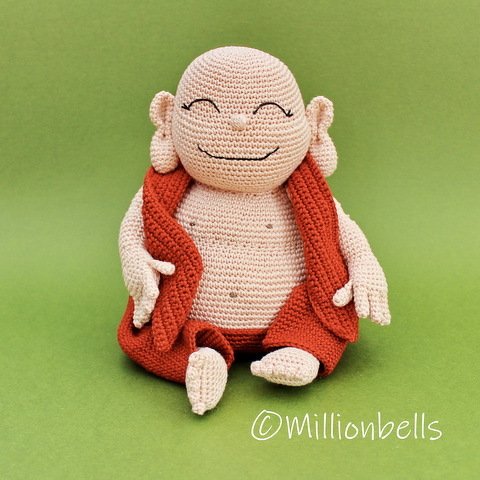 Small beige crocheted figurine with a smiling face and reddish-orange vest seated on a green surface.