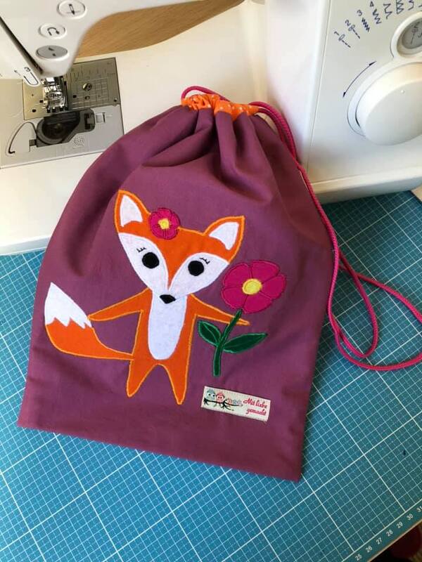 Violetter Zugbandbeutel mit aufgenähter Fuchs-Applikation und Blumenmotiv neben einer Nähmaschine auf Schneidematte.