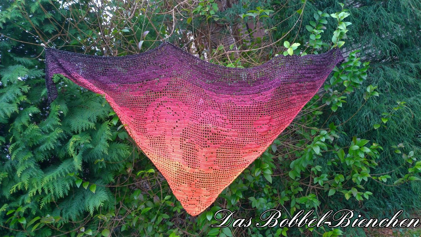 Gehäkeltes Dreieckstuch mit Rosen-Filetmuster und Farbverlauf von Violett zu Orange, aufgehängt vor grünen Hecken