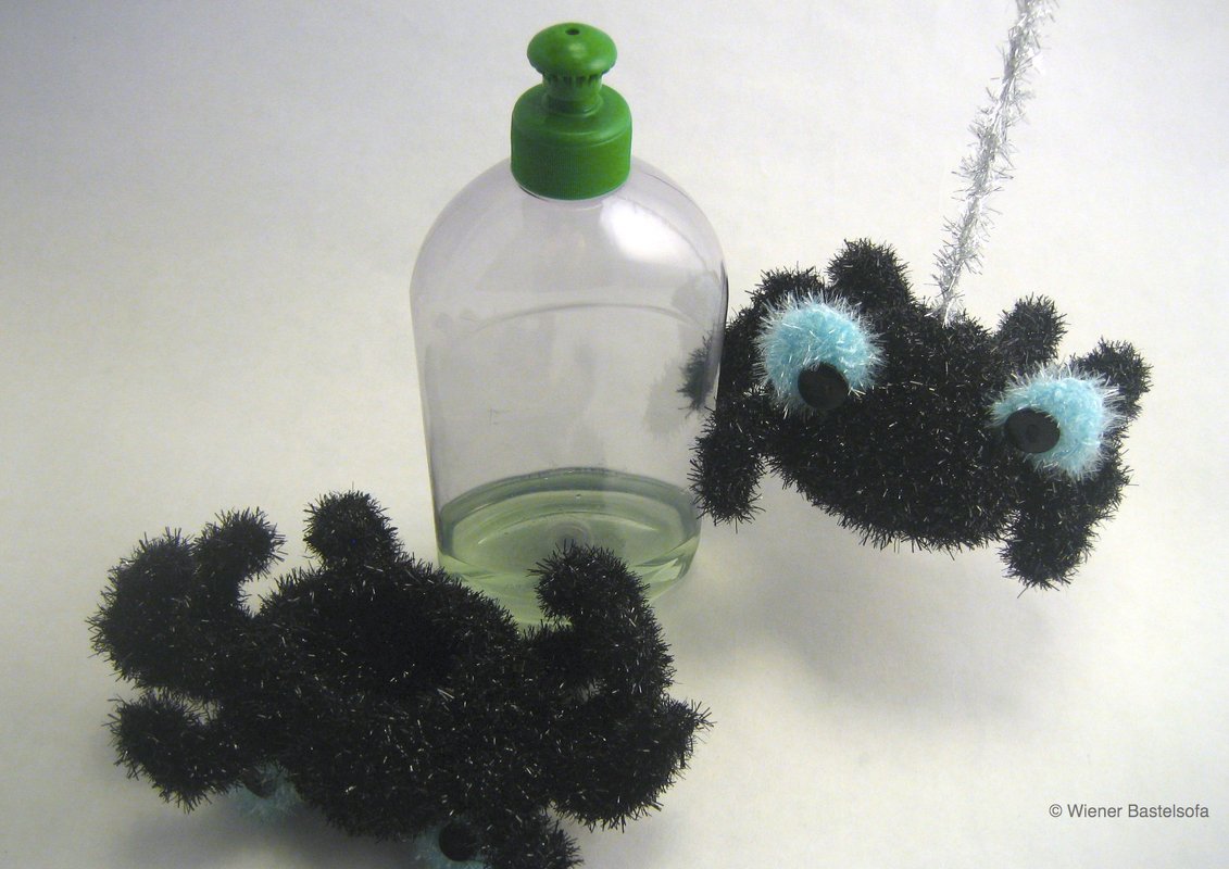 Zwei flauschige schwarze Spinnen mit hellblauen Augen und schwarzen Knopf-Akzenten; eine hängt an einem silbernen Faden, die andere liegt neben einer durchsichtigen Flasche.