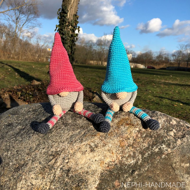 Zwei gehäkelte Wichtel mit pinker und türkisfarbener Mütze und gestreiften Beinen auf einem Felsen.