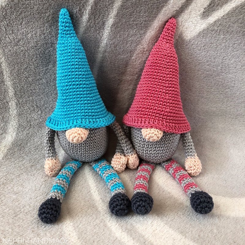 Zwei gehäkelte Amigurumi-Wichtel mit spitzen Hüten (blau und pink) auf grauer Decke