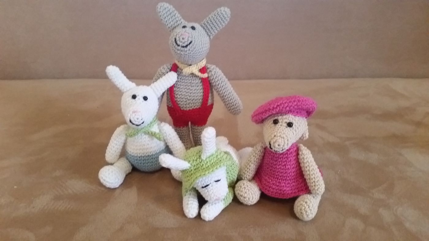 Vier gehäkelte Amigurumi-Hasen: grauer Hase mit roten Trägern, weißer mit grünem Halstuch, beiger mit rosa Mütze und ein liegender schlafender Hase mit grüner Kapuze