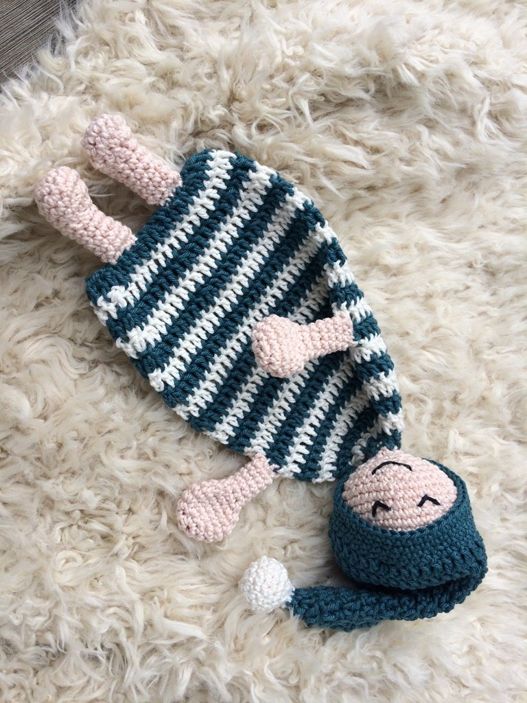 Gehäkeltes Schmusetuch in Petrol und Weiß mit zartrosa Kopf und Bommelmütze auf hellem Fell
