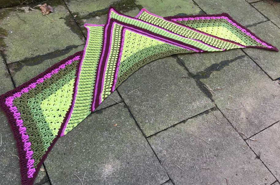 Handgehäkeltes Dreieckstuch mit breiten Streifen in Leuchtgrün, Magenta und Dunkellila auf Pflastersteinen.