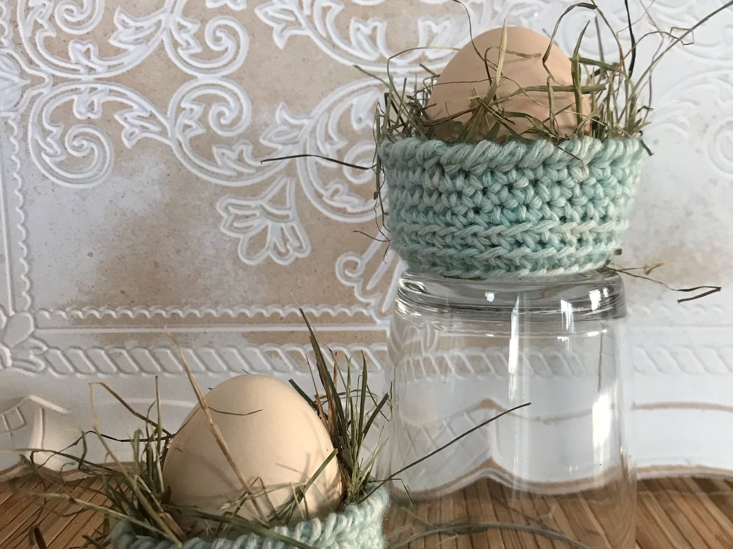 Zwei hellblaue gehäkelte Eierkörbchen, gefüllt mit Heu und braunen Eiern; eines steht auf einem Glas vor dekorativer Tapete.