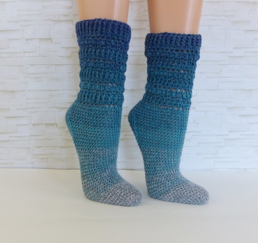 Paar gehäkelter Socken in Türkis-Grau an Beinmodellen vor heller Wand