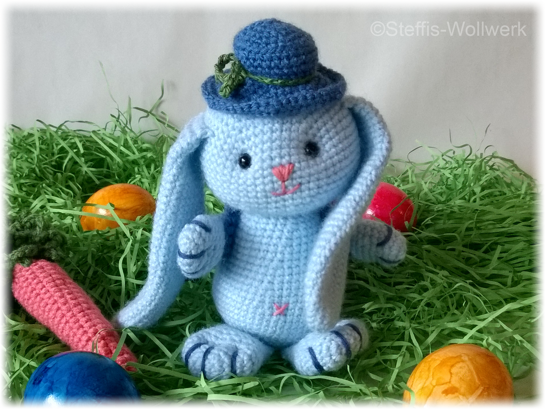Hellblauer gehäkelter Hase mit blauem Hut, Ostergras, bunten Eiern und Häkelkarotte