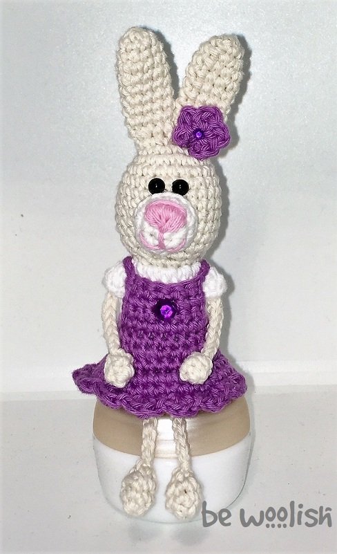 Gehäkelter Amigurumi-Hase in cremeweiß mit lila Kleid und Blume, sitzend