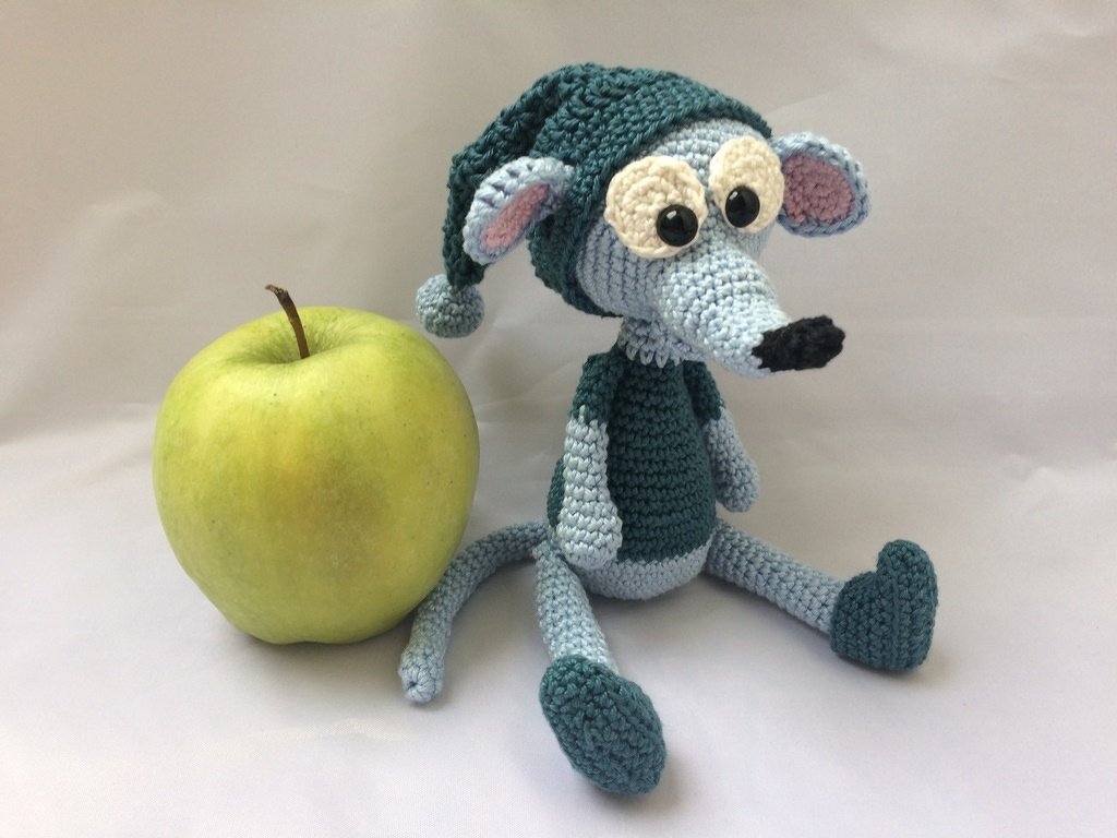 Zweifarbige gehäkelte Amigurumi-Maus in Hellblau und Petrol mit Zipfelmütze sitzt neben einem grünen Apfel auf hellem Hintergrund.